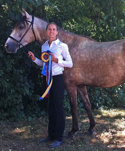 A female rider standing outside next to a tan horse and holding a prize ribbon.